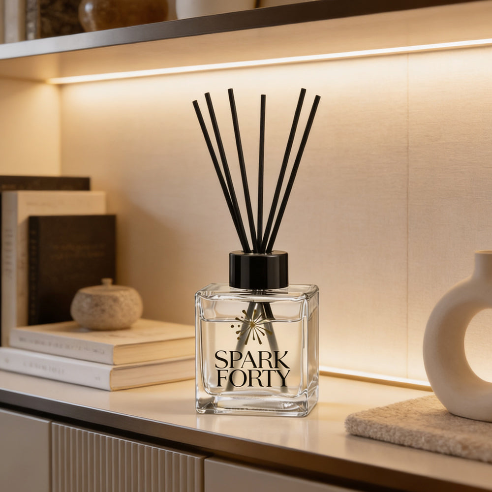 Sparkforty Rouge 540 reed diffuser displayed on a modern shelf with books and decorative accents. Luxury home fragrance diffuser in a clear glass bottle with black reeds, inspired by amber, jasmine, saffron, and woody notes for long-lasting freshness in living rooms, bedrooms, offices, and entryways.